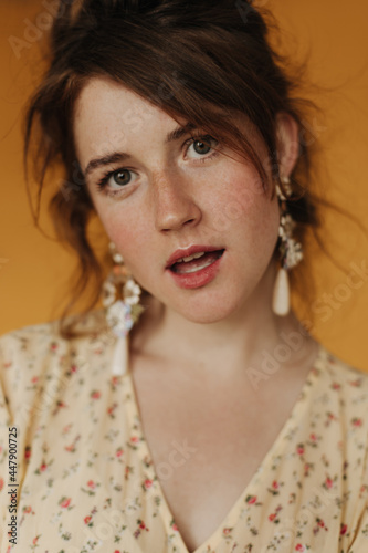 Portrait photo of stylish girl with freckles looking into camera on isolated backdrop. Charming lady in flower outfit posing on yellow background..