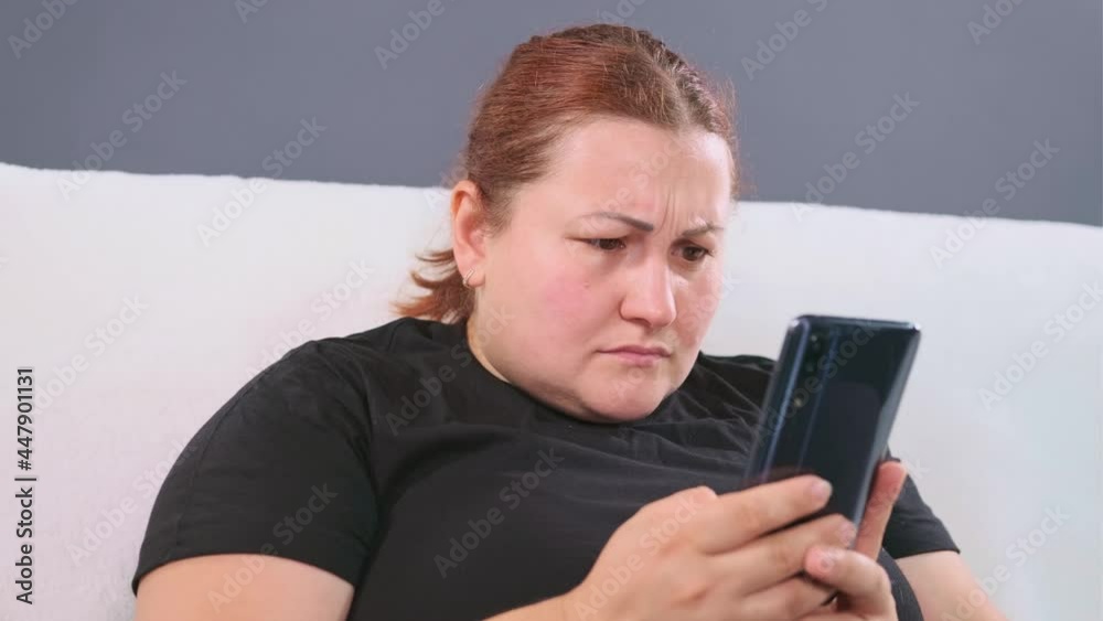 Woman sitting on the white sofa holding the mobile phone in her arms and viewing emotionally news on the internet, having worried facial expression. Woman sitting on the white sofa holding the mobile phone in her arms and viewing emotionally news on the internet, having worried facial expression.