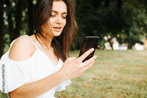 Young brunette woman in white summer dress using mobile phone, chatting on smartphone in social media, browsing the internet outdoors in the park. Selective focus on the mobile phone.