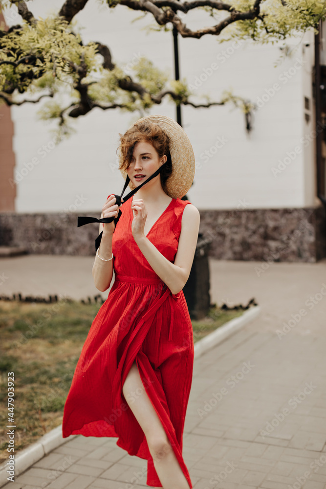 Obraz premium Bright girl in red summer dress posing at street. Beautiful woman with brown hair putting modern hat and looking away outdoors..