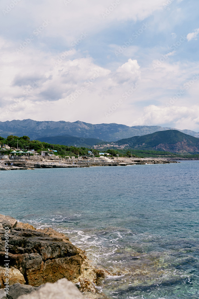Sea waves hit the rocky shore against the backdrop of mountains, greenery and buildings