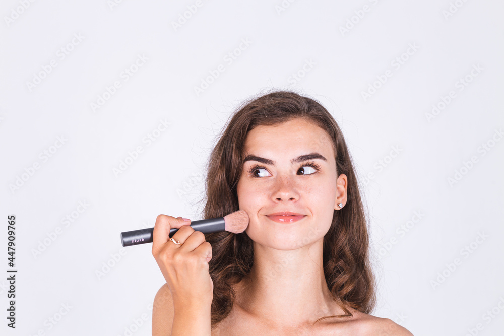 Young beautiful natural soft clean skin woman with freckles light makeup on white background with bare shoulders holding blush brush Young beautiful natural soft clean skin woman with freckles light makeup on white background with bare shoulders holding blush brush