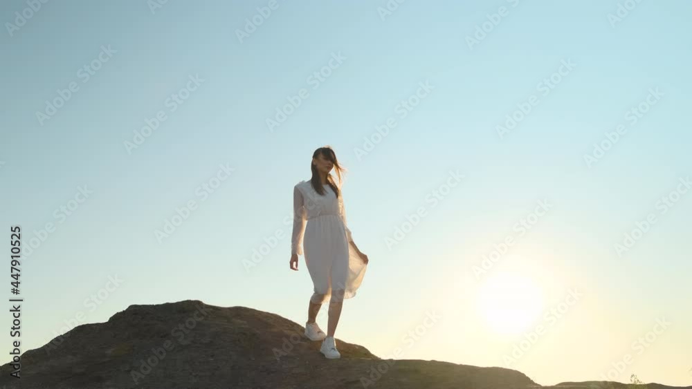 beautiful slender woman in translucent white dress and white sneakers descending from top of hill, sun shines into camera, sunset or dawn. young lady walks through mountains with sky in background beautiful slender woman in translucent white dress and white sneakers descending from top of hill, sun shines into camera, sunset or dawn. young lady walks through mountains with sky in background