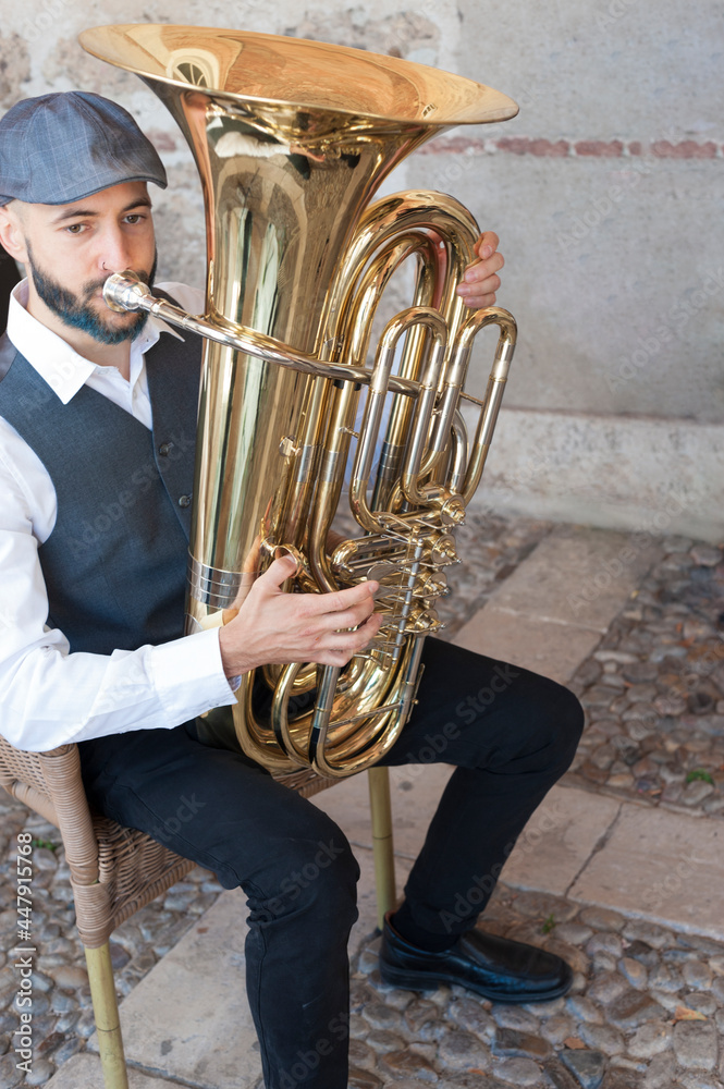 Foto Stock A jazz musician dressed in vintage style, playing the tuba on the  street in broad daylight. With beard and goatee dyed blue and a retro cap.  | Adobe Stock