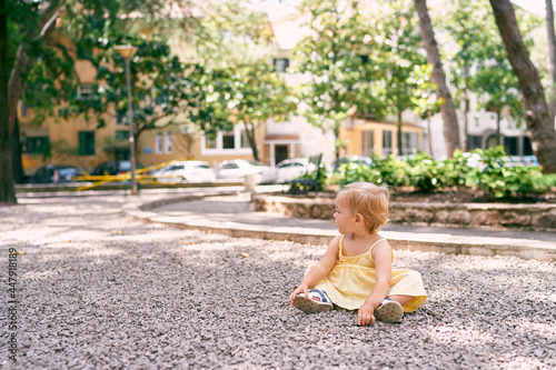 Little girl in a dress sits on a gravel path in the courtyard of the house