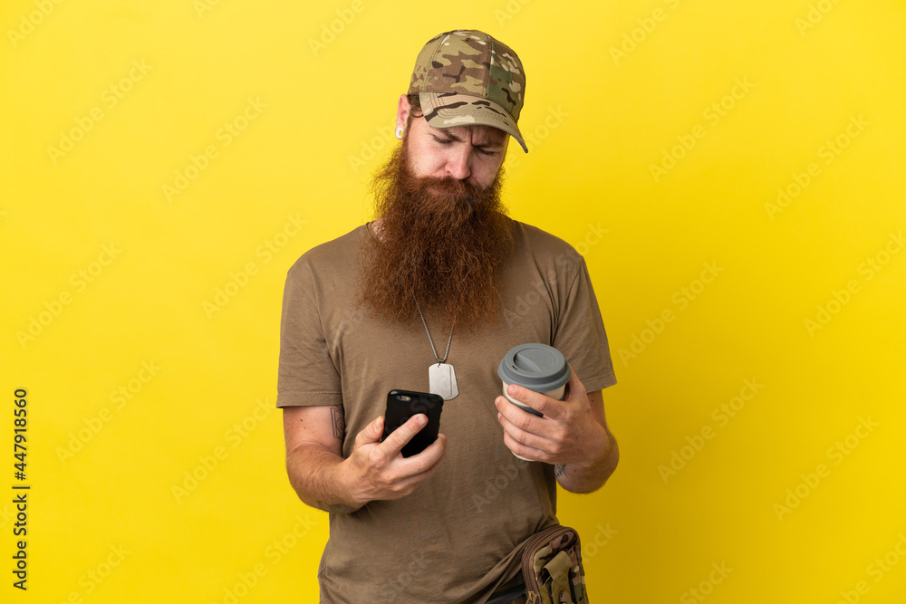 Fototapeta premium Redhead Military man with dog tag isolated on yellow background holding coffee to take away and a mobile