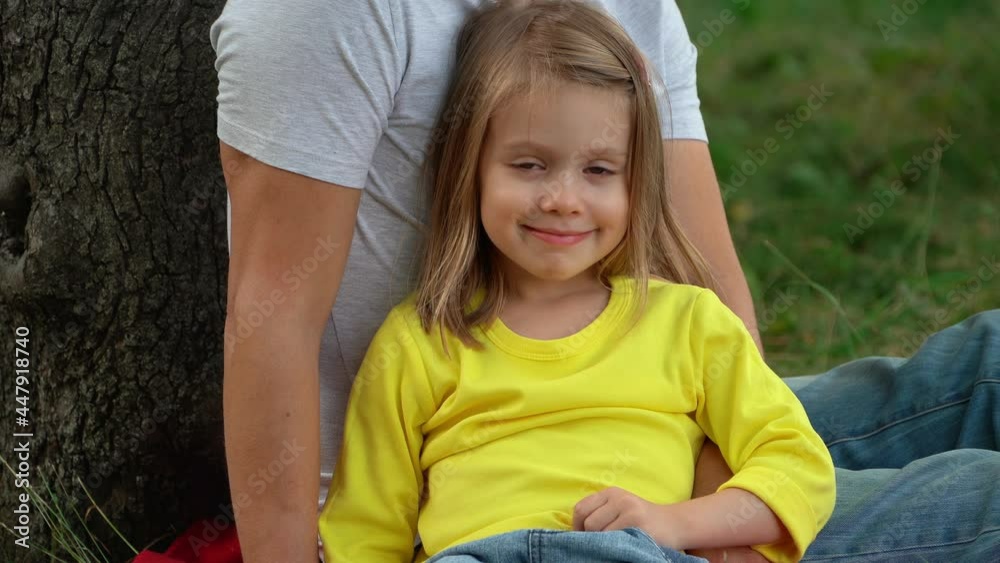 Portrait of little girl sits next to tree on green lawn in summer park Happy child looks at camera and smiles. Outdoor picnic and recreation concept Portrait of little girl sits next to tree on green lawn in summer park Happy child looks at camera and smiles. Outdoor picnic and recreation concept