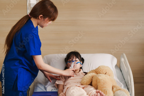 Young Asian female caregiver holding oxygen mask with cute girl patient in hospital. Nurse putting putting inhalation on the girl face. photo