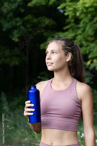 Vertical photo of a young athlete with a bottle of water in the forest on a blurry background of trees. A sports girl stands with a bottle in nature in the park in the fresh air.