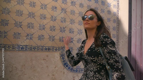 young happy caucasian tourist in glasses with backpack travels in portugal.girl walks down the street and touches traditional blue azulejo tiles on an old building in lisbon. woman enjoys architecture photo