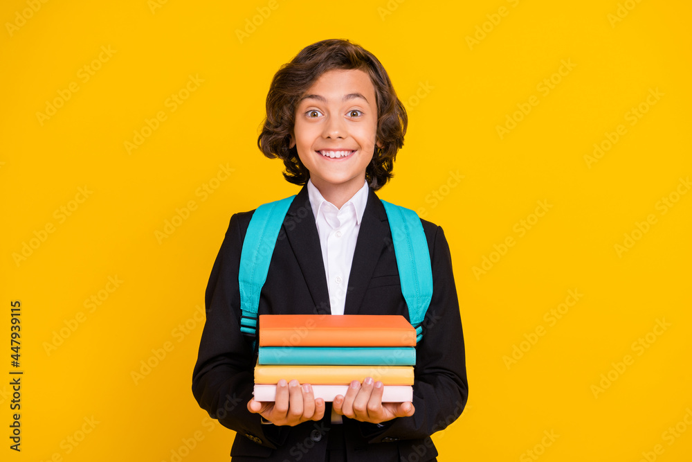 Photo of positive funny little schoolboy hold pile book wear rucksack black uniform isolated yellow color background Photo of positive funny little schoolboy hold pile book wear rucksack black uniform isolated yellow color background