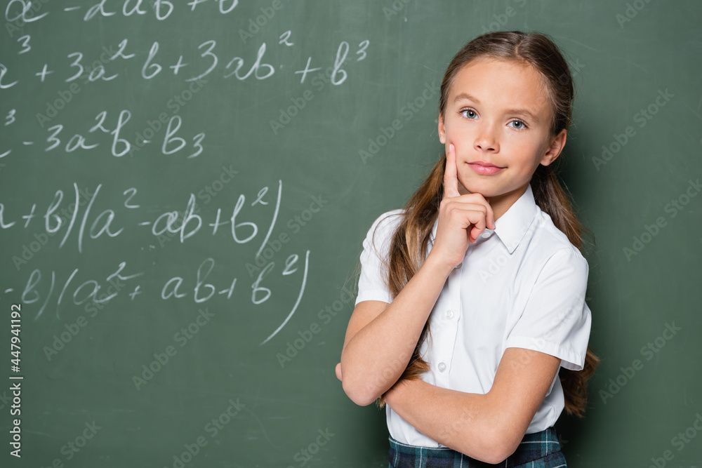 pensive schoolgirl with hand near face looking at camera near chalkboard with equations pensive schoolgirl with hand near face looking at camera near chalkboard with equations