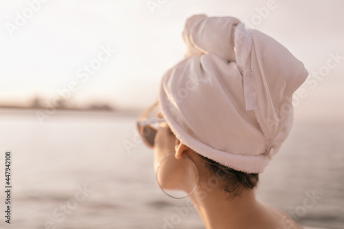 Young lady with hair wrapped in towel, stylish earrings and sunglasses looking at sea and enjoying pastel pink sunset