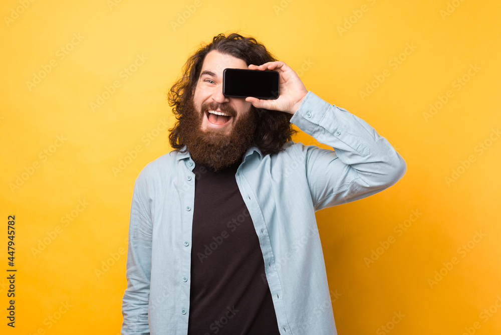 Joyful young bearded man with long hair holding smartphone over eye and smiling over yellow background Joyful young bearded man with long hair holding smartphone over eye and smiling over yellow background
