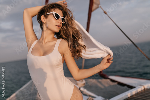 Well-built young blonde in white swimsuit, trendy sunglasses and round earrings standing on yacht and looking away against dark blue sea background © Look!