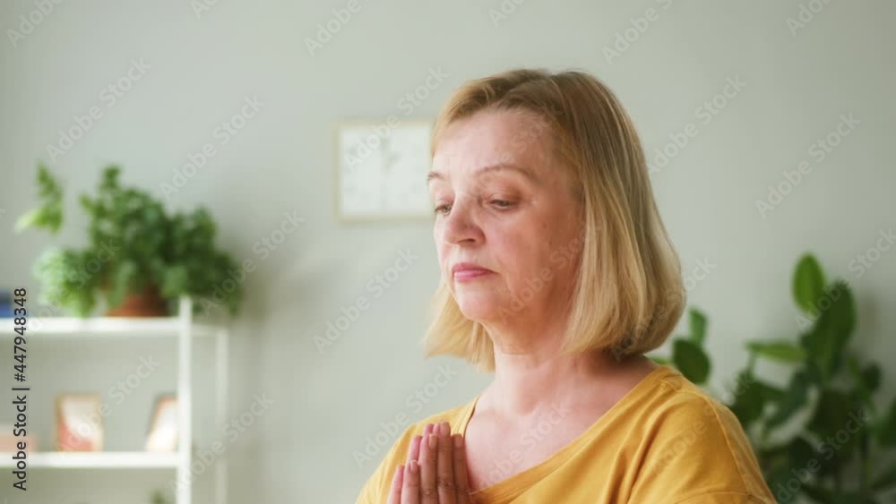 Elderly woman wearing sports clothes sitting in lotus position on gym mat in living room. Female person doing yoga on floor at home, relaxing and meditating in morning. Healthy lifestyle and long life Elderly woman wearing sports clothes sitting in lotus position on gym mat in living room. Female person doing yoga on floor at home, relaxing and meditating in morning. Healthy lifestyle and long life