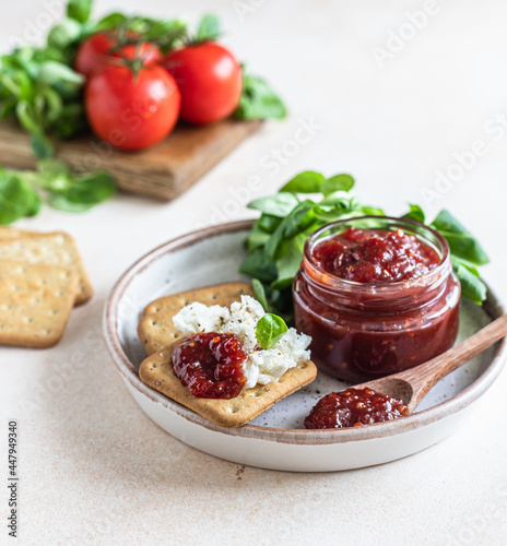 Tomato jam, confiture or sauce in glass jar with crackers and green leaves salad. Unusual savory jam. Mediterranean cuisine.