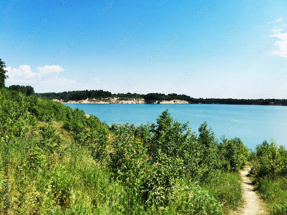Beautiful blue lake surrounded by forest. Grass and flowers by the lake in a sandy quarry