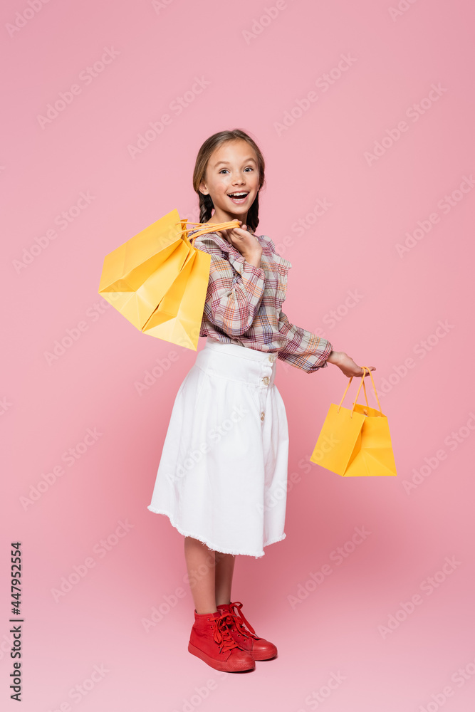 amazed kid in plaid blouse and white skirt holding yellow shopping bags on pink background amazed kid in plaid blouse and white skirt holding yellow shopping bags on pink background