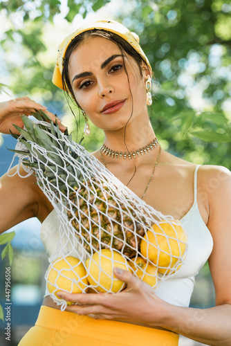 trendy woman in yellow headscarf holding string bag with fruits