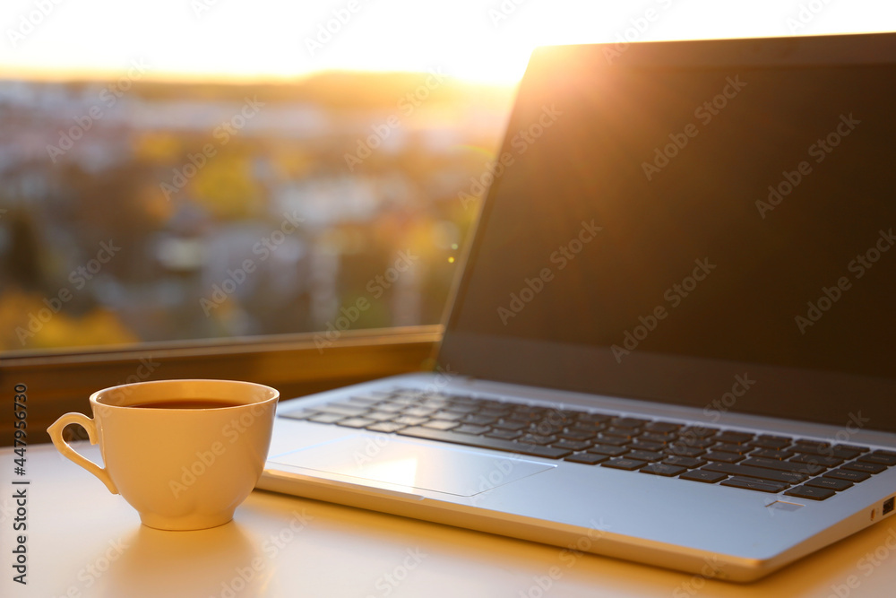 Fototapeta premium Hot coffee cup and laptop on table against the window at sunrise. Cozy workplace in home office, concept of remote work and earning online