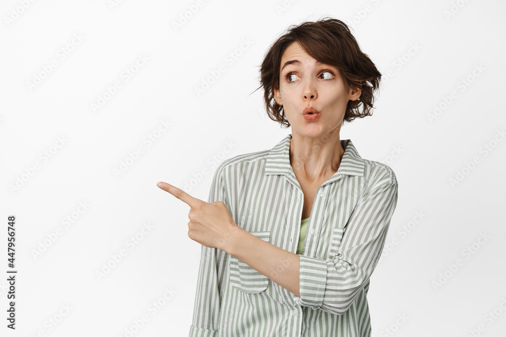 Portrait of surprised young woman pucker lips curious, pointing and looking left at interesting promo, advertisement, standing against white background Portrait of surprised young woman pucker lips curious, pointing and looking left at interesting promo, advertisement, standing against white background