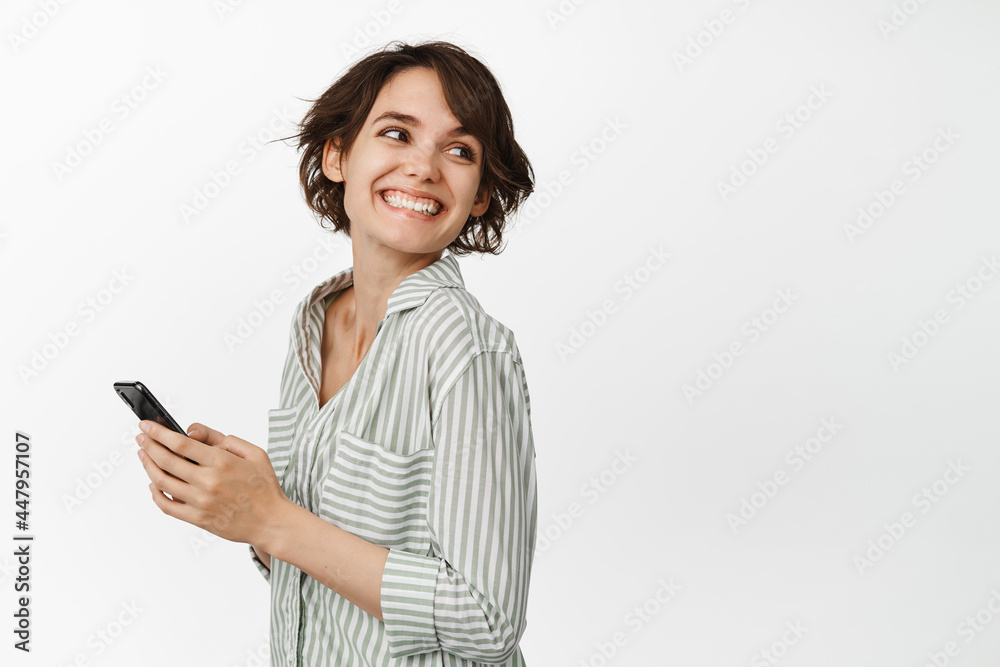 Portrait of young smiling woman holding smartphone, turn head at copy space, chatting cellular, standing against white background Portrait of young smiling woman holding smartphone, turn head at copy space, chatting cellular, standing against white background