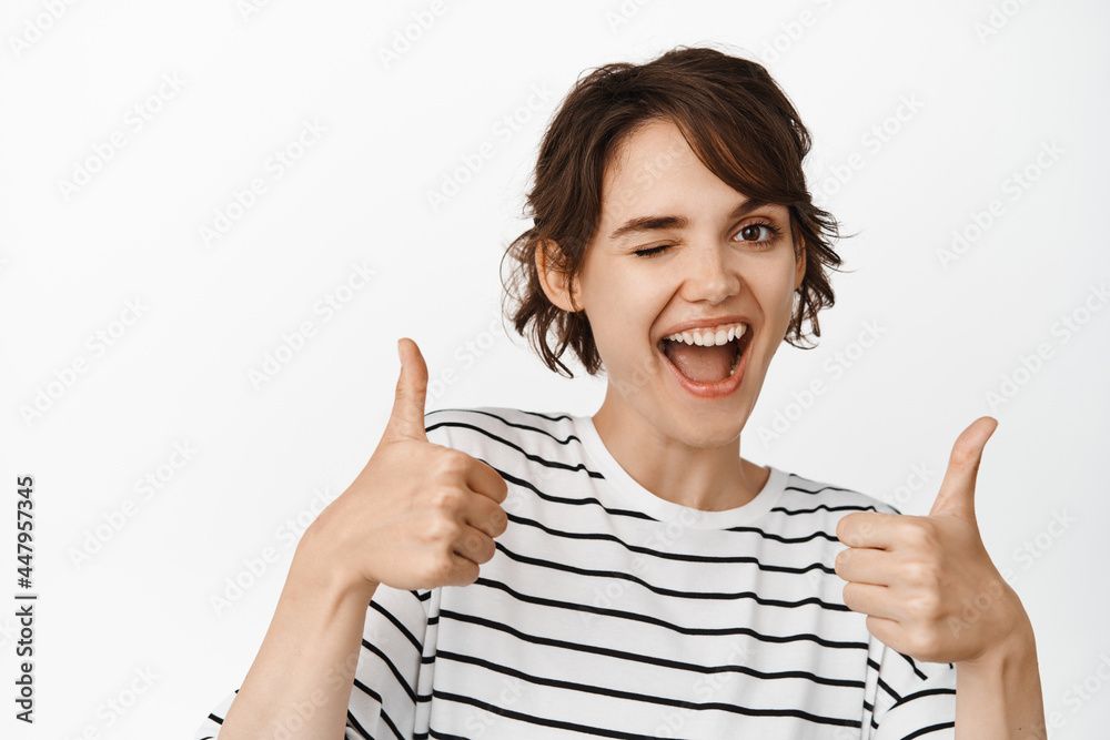 Good job, excellent choice. Smiling brunette girl showing thumbs up and winking, look pleased, standing over white background Good job, excellent choice. Smiling brunette girl showing thumbs up and winking, look pleased, standing over white background