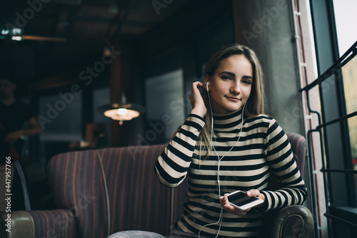 Portrait of Caucaisan millennial blogger in electronic earphones looking at camera, beautiful hipster girl with modern mobile gadget in hand posing while listening audio book in cafe interior