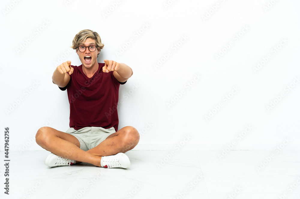 Young English man sitting on the floor surprised and pointing front Young English man sitting on the floor surprised and pointing front