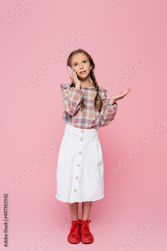 full length view of stylish child gesturing while talking on cellphone on pink background