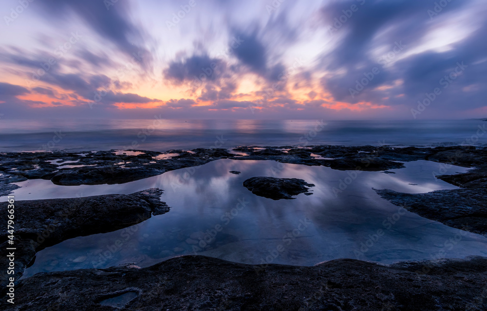 Naklejka premium amazing scenic view at a sea shore from a coast after dramatic colorful sunset with flying blured clouds and reflection on water surface , beautiful ocean landscape