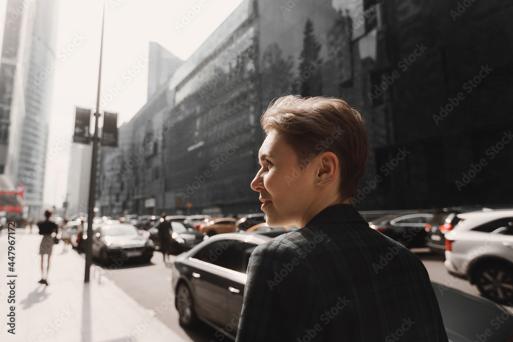 beautiful modern business woman with a short haircut and a cloaky jacket walking along the road between buildings against the background of cars. Sunlight.