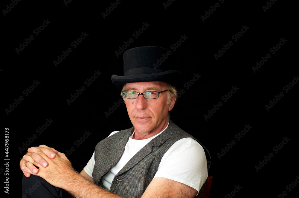 Studio portrait on caucasian man wearing top hat, vest and glasses, sitting with hands on knee looking at viewer. Studio portrait on caucasian man wearing top hat, vest and glasses, sitting with hands on knee looking at viewer.