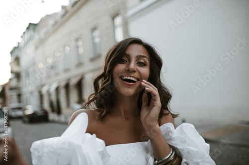 Portrait of joyful young brunette with wavy hair, tanned body in white vintage top and accessories smiling and looking into camera against day city background 