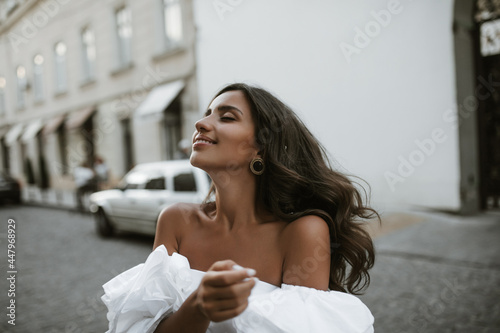 Charming young brunette with bronzed body in white off-shoulder blouse and vintage accessories  smiling and posing with closed eyes on daylight street