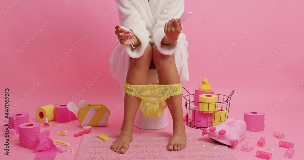 Menstruation and periods concept. Unknown woman in white bathrobe holds sanitary napkin cotton tampon chooses best alternative sits on toilet bowl surrounded by paper rollers feminine items.