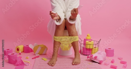 Menstruation and periods concept. Unknown woman in white bathrobe holds sanitary napkin cotton tampon chooses best alternative sits on toilet bowl surrounded by paper rollers feminine items.