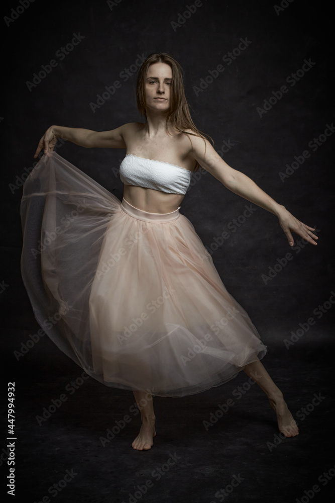 Fototapeta premium Young girl in white top and peach skirt dancing in black old textured studio