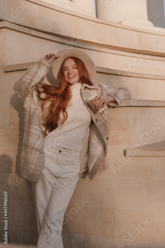 Happy young foxy lady standing against architecture background outdoors. Adorable long-haired girl with light clothes, plaid coat, pants and stylish hat, holding cup of coffee in hand and smiling