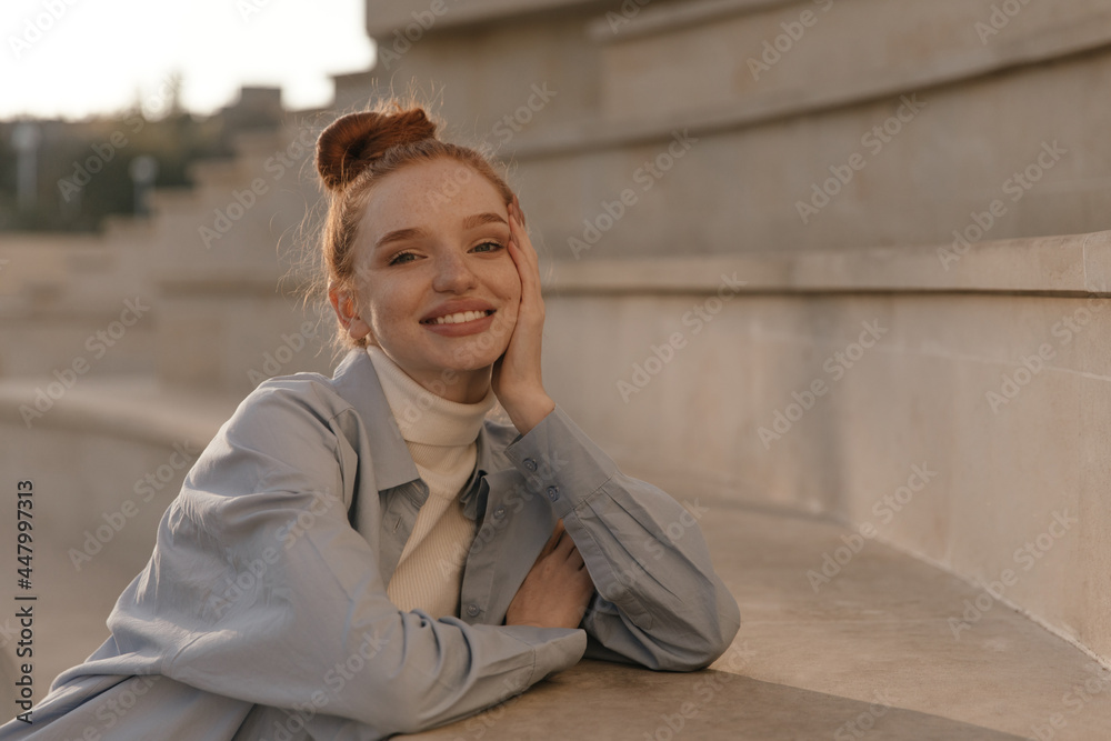 Fototapeta premium Frontal portrait of joyful young ginger chilling at sunny city. Adorable lady with collected hair and light clothes, smiling, looking into camera and posing outdoors