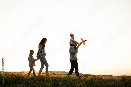 Parents with children walk in the summer at sunset walking across the field. Dad is carrying a baby with a airplane on his shoulders. Mom leads her daughter by the hand. Foster family.