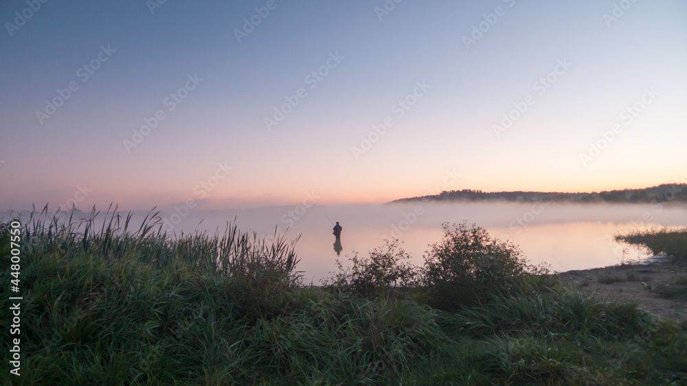 A fisherman stands knee-deep in water and catches fish on a foggy autumn morning