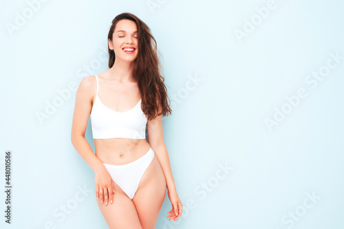 Portrait of young beautiful smiling woman in white lingerie. Sexy carefree cheerful model in underwear posing near light blue wall in studio. Positive and happy female enjoying morning © halayalex