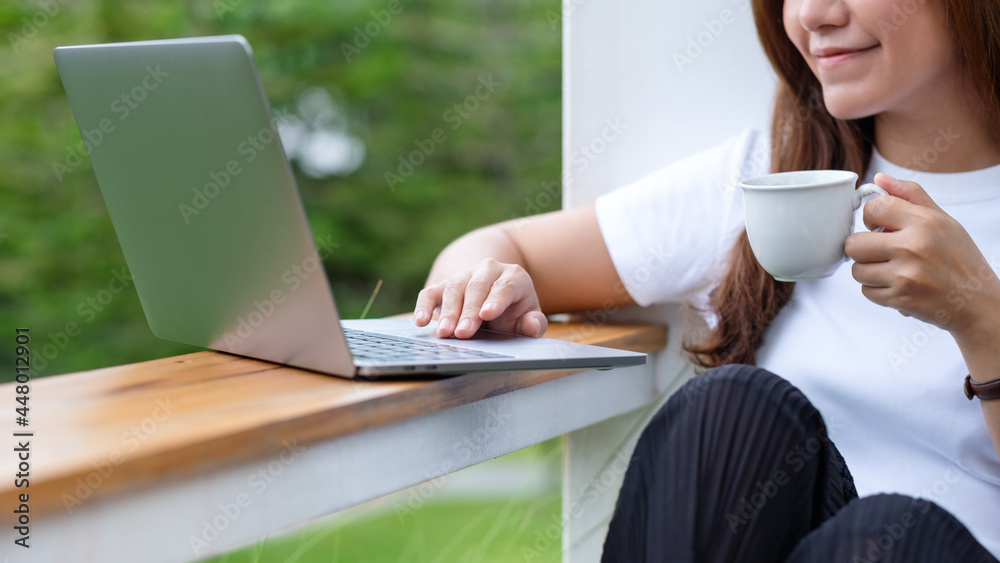 Closeup image of a beautiful young asian woman drinking coffee while using and working on laptop computer in the outdoors Closeup image of a beautiful young asian woman drinking coffee while using and working on laptop computer in the outdoors