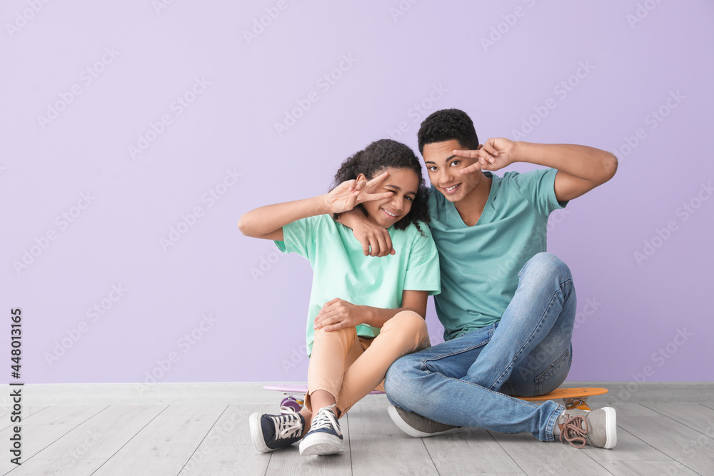 African-American brother and sister with skateboards showing victory gesture while sitting near color wall