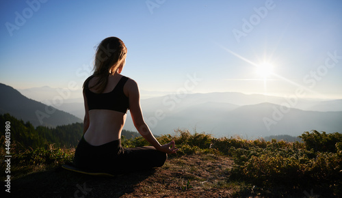 Back view of brunette practicing yoga in Lotus pose. Female is meditating on grass outdoor and watching sunset.