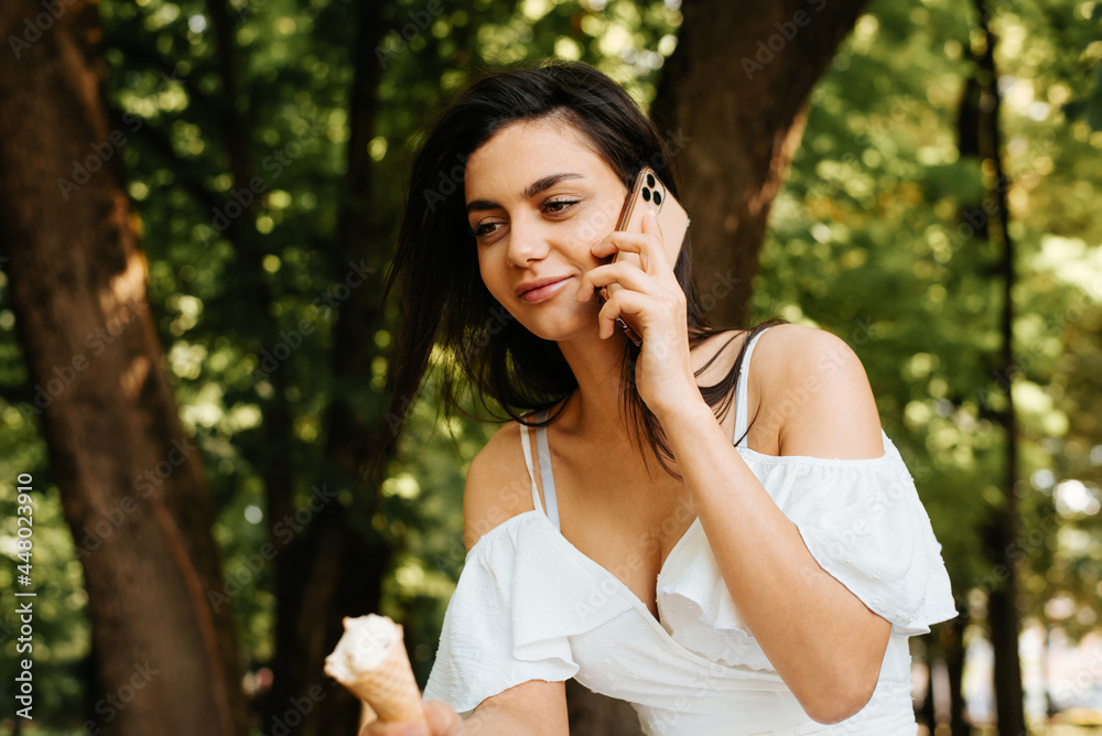 Portrait of a beautiful young Indian woman talking on a cell phone and holding an ice cream, outdoors. Smiling brunette woman in white dress using smartphone, lifestyle. Portrait of a beautiful young Indian woman talking on a cell phone and holding an ice cream, outdoors. Smiling brunette woman in white dress using smartphone, lifestyle.