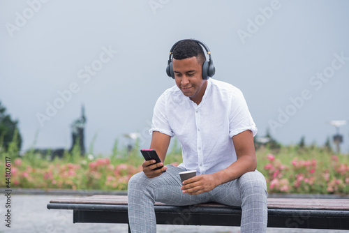 Portrait of a young and happy African American man with headphones. A man sitting on a bench and listening to music, holding a smartphone and a mug of coffee © Павел Костенко
