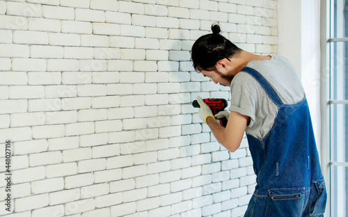 Young Asian man using electric drill on white brick wall in room, concept for home improvment, DIY work, do it youself photo